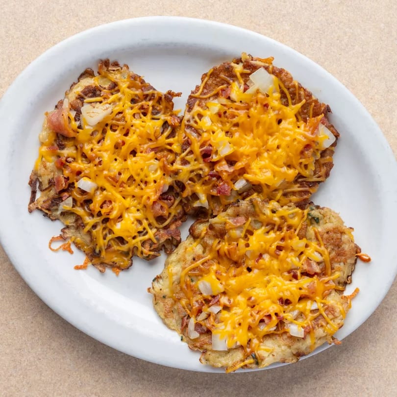 Three golden-brown hash browns topped with melted cheddar cheese, bacon bits, and diced onions on a white plate.
