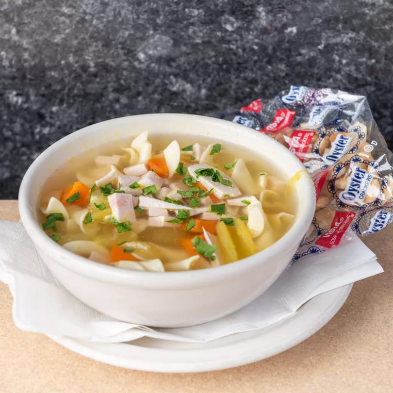 A white bowl of chicken noodle soup with carrots and parsley, served with oyster crackers.