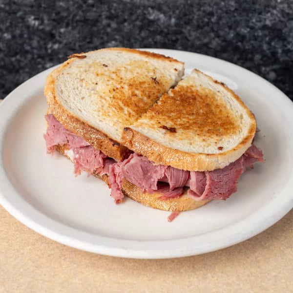 A toasted pastrami sandwich on a white plate, with a dark countertop in the background.